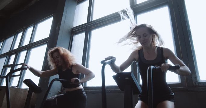 Two young smiling athletic European women work out on gym bicycle machines during cardio training, slow motion.