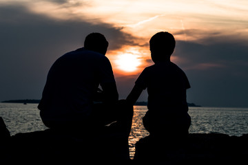 Father And Son Talking While Sitting On Stones At The Coast During Sunset