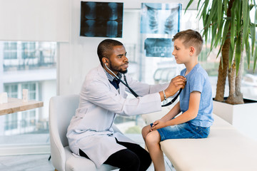 Young smiling African American male doctor examining little boy in modern clinic