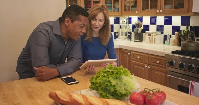 Mature African American And Caucasian Couple Standing In Kitchen Looking At Recipe On Tablet Computer. Husband And Wife Cooking Together Checking Online For Instructions. Slow Motion 4k Handheld