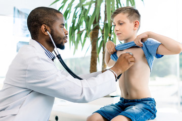 African male doctor and child boy patient. Smiling pediatrician man sees boy in medical office. African American male pediatrician with stethoscope listening to lung and heart sound of little boy © sofiko14