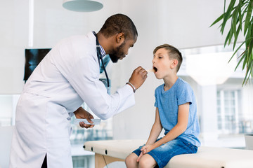 Obraz premium Showing throat. Little boy opening mouth showing her throat while visiting young handsome African man doctor in his medical office