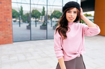 Image of young brunette woman smiling and looking to the camera in the city. Modern stylish female wearing pink sweater and black hat beret against building wall in the city street. Natural lighting