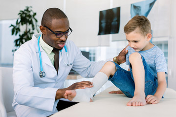 Leg injured boy visiting young African doctor traumatologist in his medical clinic hospital. Boy is sitting on the couch. Doctor in white coat examines the leg