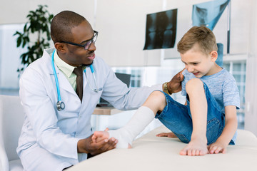 Fototapeta premium African-American pediatrician examining boy in clinic with leg sprain with elastic bandage.