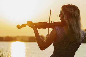 romantic silhouette of young woman with a violin at dawn on river bank, elegant girl playing a...