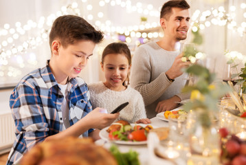 technology, holidays and people concept - happy children with smartphone at family dinner party
