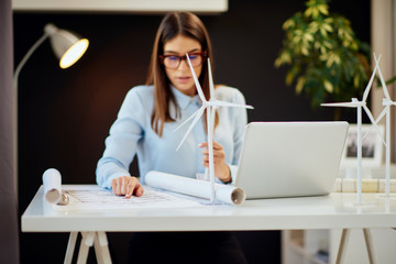 Charming hardworking brunette dressed in formal wear sitting at desk in office, holding windmill model and looking at plans. On desk is laptop. Selective focus on windmil models.