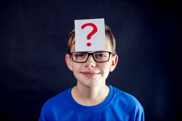 A portrait of a boy with eyeglasses and question mark at his head in front of dark background. Children and education concept