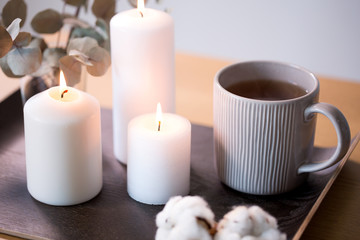 Obraz na płótnie Canvas decoration, hygge and cosiness concept - burning white candles, tea in mug, branches of eucalyptus populus and cotton flowers on table