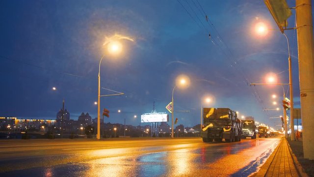 A convoy of military equipment rides through the city at night with headlights on