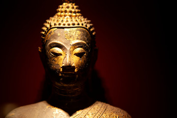 Close up. Golden Buddha metal statue with closed eyes in a dark place with light above it. Meditation process. Asian culture in Milan, Lombardy, Italy.