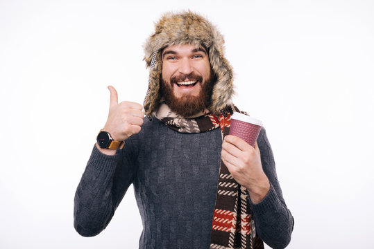 Good Coffee Joyful bearded hipster wearing scarf and winter hat showing like gesture while holding a takeaway cup.