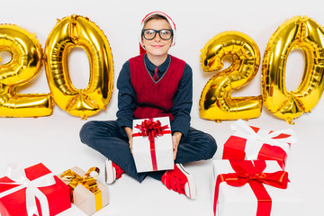 Happy little boy in Santa hat, stylish baby with Christmas gifts, sits on white background with...