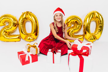 Happy little girl in Santa hat, stylish baby sitting with Christmas gifts sitting on white...