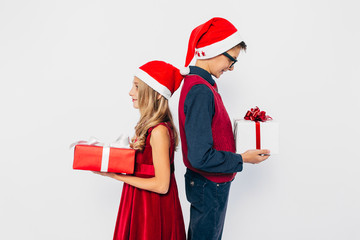 little girl and boy in Santa hat, stylish brother and sister, make each other surprise by holding Christmas gifts in hands while standing on white background