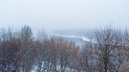 Winter landscape - river in the fog, bare tree branches on the slope of a high bank