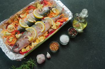 Baked fish with vegetables and lemon and spices in a baking dish. Top view on a black background. Copy space