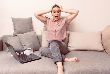 1 white young woman in a pink blouse and gray jeans sits on a couch with a laptop and a document and screams in terror, holding her head with her hands,