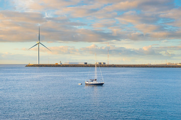 Coastal landscape with calm bay, breakwater, windmill and yacht at sunset