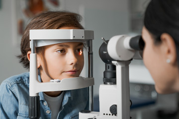 Fototapeta na wymiar Adult female doctor ophthalmologist is checking the eye vision of cute child in modern clinic