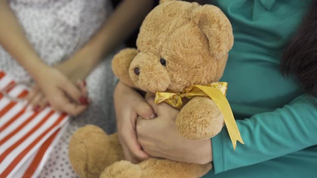 Close-up of soft yellow teddy bear with a silk bow. Female hands holding the gift. Pregnant woman in silk blue-green dress with a present for her unborn child.