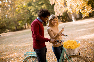 Multiracial couple with bicycle standing in the autumn park and using mobile phone