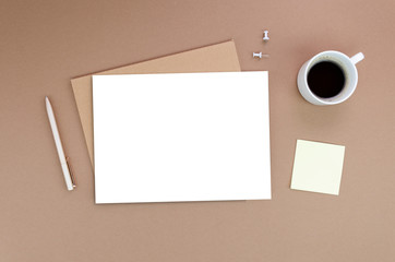 Top view of a desk mockup: craft envelope with a blank paper list and a white pen, coffee cup on a side on a beige background