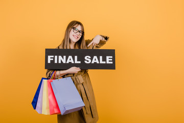 happy young woman in fall trench coat and glasses with final sale sign and colorful shopping bags isolated over yellow