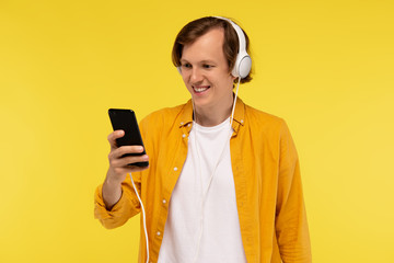 smiling guy listen popular song in headphones, smile broadly, feel rebellious and using smartphone isolated on yellow background. Blogger concep