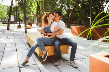 A young couple is sitting on a bench in a modern city, hugging and smiling.