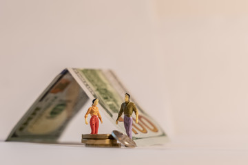 Miniature woman and man figure standing next to house made of dollar bill and counting coins. Shallow depth of field background. Family budget, mortgage and cost of housing concept.