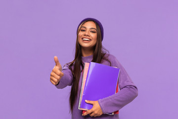 Smiling african-american teenager student in a hat and sweater holding folders and showing thumb up...