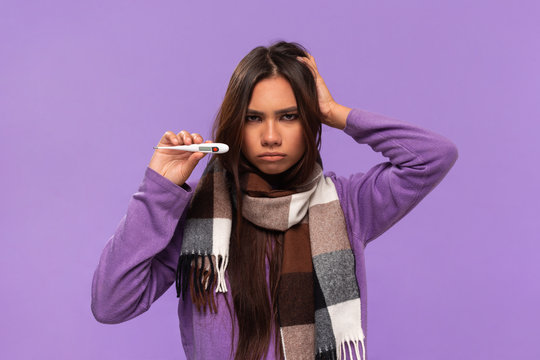 Pretty african-american girl in a sweater and cosy checked scarf checking thermometer standing isolated over purple background. Catching cold in winter.