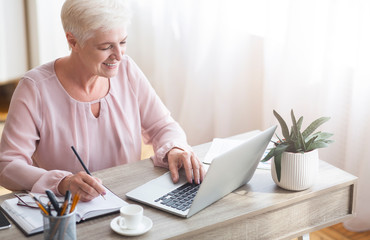 Fototapeta premium Top view of mature working lady making notes, using laptop