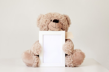 Mockup. Soft beige teddy bear toy holding white clean mock up frame with copy space sitting at light grey background. Empty space. Baby children concept.