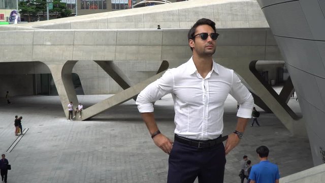 Mid shot of handsome and fit European man with dark hair in white collared shirt is standing on a grand staircase in modern setting waiting for someone. He puts on his sunglasses and looks around