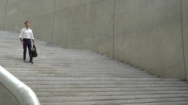 Mid shot of handsome and fit European man with dark hair in white collared shirt and dark trousers is walking down a grand modern concrete staircase towards camera and out of frame