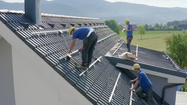 Three workers in blue shirts and yellow helmets doing cable instalation for solar panels on the roof - steady, closup drone shot with a view from a side