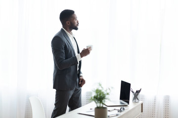 Pensive businessman standing near window in office and drinking coffee