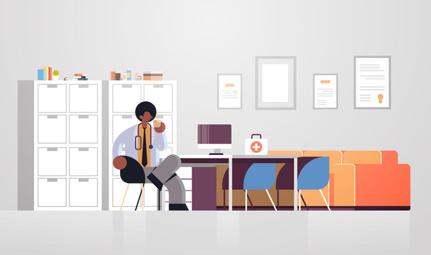 male doctor in white coat having coffee break medicine healthcare concept african american medical worker sitting at workplace modern hospital clinic office interior full length flat horizontal