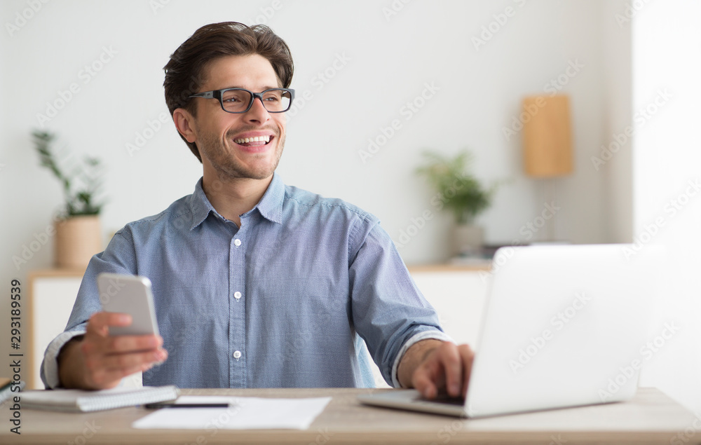 Poster man using smartphone sitting in front of laptop in office