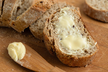 Closeup of buttered slice of whole wheat rustic bread next to cut bread and wood knife. Light wood board.