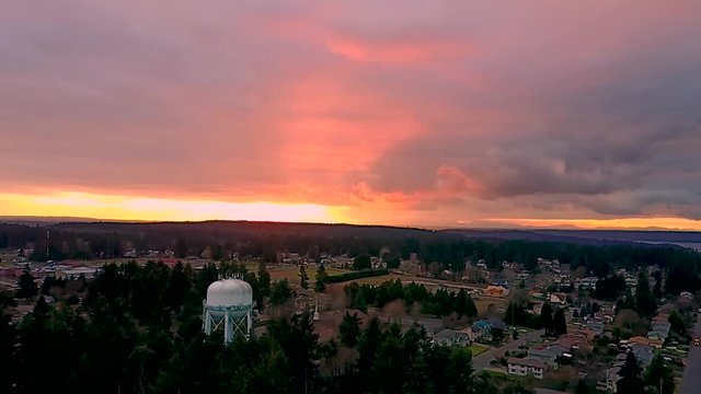 Wide aerial shot with forward movement showing a cloudy, orange sunset and a water tower overlooking a small town in the trees in Steilacoom, Washington.