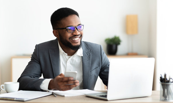 Portrait of smiling black businessman sitting at workplace with cellphone