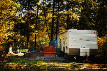 A deserted camper trailer and tent with wooden chairs on outdoor carpet and cloth covered table surrounded by tall pine trees parked in a deserted campsite in a autumn daytime landscape