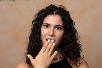 A beautiful twenty year old girl is seen gasping in shock as she holds hand over her open mouth with white lacquered nails and wavy brunette hair, standing against a neutral background.