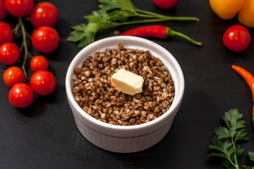 Cooked buckwheat porridge in a deep plate on a black wooden background with vegetables.