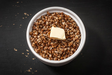 Cooked buckwheat porridge in a deep plate with a slice of butter on a black wooden background.