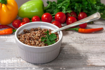 Cooked buckwheat porridge in a deep plate wiht spoon on a light white wooden background with vegetables